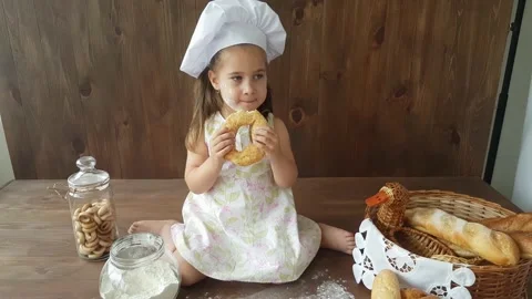A three-year-old girl in a white dress and a baking cap is eating a big bagel Stock Footage 157126441