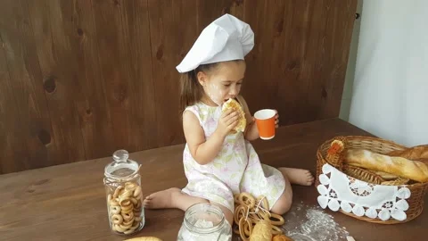 A three-year-old girl in a white dress  eats a large bagel and drinks with juice Stock Footage 157126467