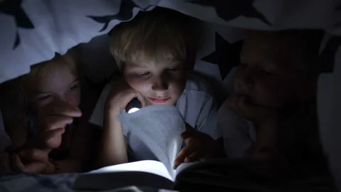 Three young children are reading a book with a flashlight under the covers at Stock Footage 134863544
