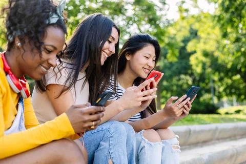 Three young multiracial girls using mobile phone sitting outside Stock Photos