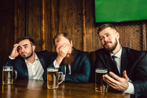 Three young office workers in suits sit at table in bar. They watch football 写真素材