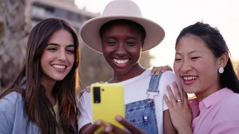 Three young smiling multi-ethnic girls using cell phone together outdoors. Group Stock Footage 250863098