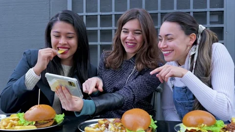 Three young women sitting at table eating burger and chips with smartphone. Video stock 166302911
