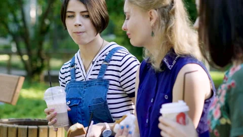 Three young women talking, drinking coffee on a wood bench in a summer park. 4K 库存影片 87724812