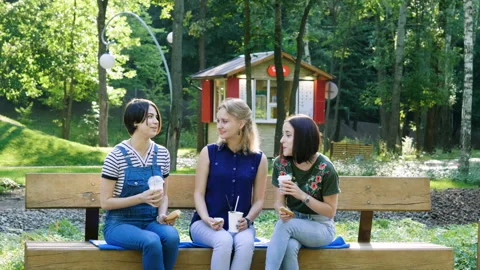 Three young women talking, drinking coffee on a wood bench in a summer park. 4K 스톡 동영상 87725139