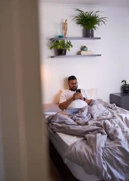 A through the door shot of a man lazing in bed on his phone Stock Photos
