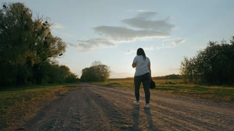 Through the rays of the setting sun, a lonely girl walks along a dirt road. Stock Footage 243119019