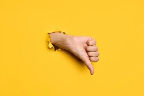 Thumb down sign gesture through hole in torn yellow paper background Stock Photos