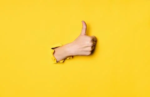 Thumb up hand gesture through hole in torn yellow paper background Stock-Fotos