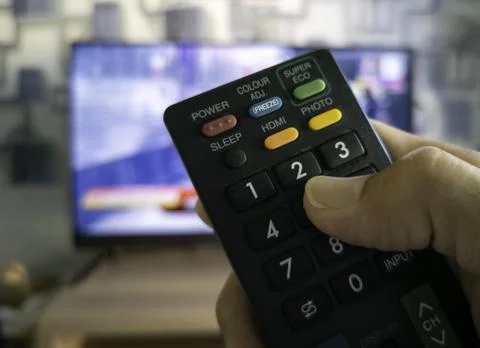 The thumb is on the remote control. Behind is a flat screen TV. Fotos de archivo