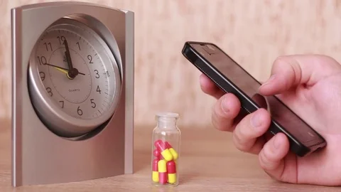 Thumbing through the smartphone in front of pills and clock Видео 83030575
