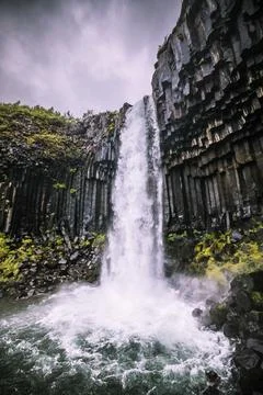 Thunderous masses of water cascade down the Skaftafell waterfall along gray c Stock Photos
