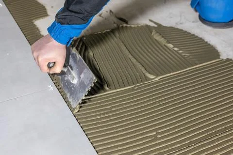 Tiler worker placing or tiling gray ceramic tile in the position over adhesive 库存照片