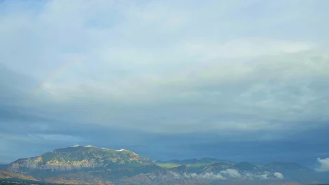 Tilt down from cloudy sky to feature rainbow over city with mountains behind Stock Footage 127559892