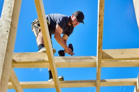 Timber frame assembly as young construction worker installs lumber at roof .. Stock Photos