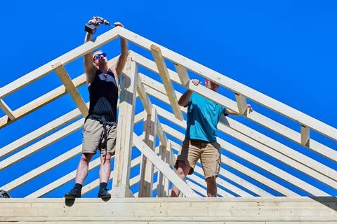 Timber trusses are assembled as two construction workers fasten rafter legs.. Stock Photos