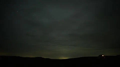 Timelapse Dark, heavy clouds roll over a tiny, glowing cabin under a deep purple Stock Footage 340240257
