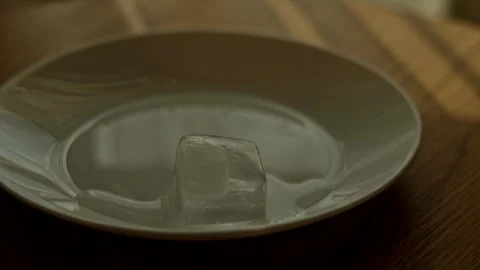 Timelapse of melting ice cube on white plate with sunlight rays moving acros Vidéo 346305216