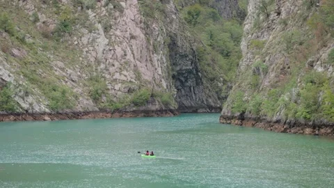 A tiny green boat navigates through calm waters bordered by steep rocky cliffs. 库存影片 336624440