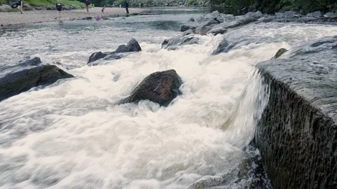 A tiny low waterfall near a metal bridge; people walking and relaxing on Stock Footage 110257133