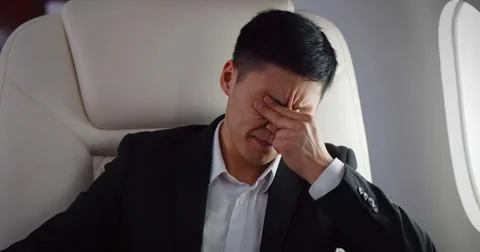 Tired asian businessman rubbing eyes sitting near window in private jet Stock Photos