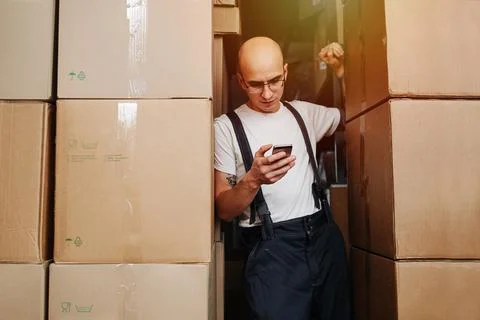 Tired bold man standing between high stacks of boxes, looking at his phone Stock Photos