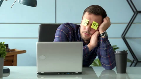 Tired comic male manager pretends working sleeping with stickers on face sits at Stock Footage 144840923