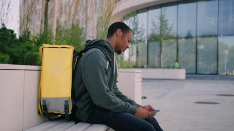 Tired courier with a backpack sits on a bench at the end of the working day Stock Footage 254098498