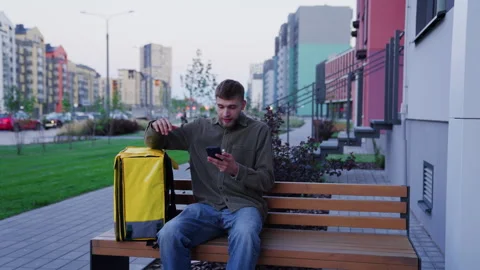 A tired courier sits down on a bench, looks next delivery on the phone Видео 265063322