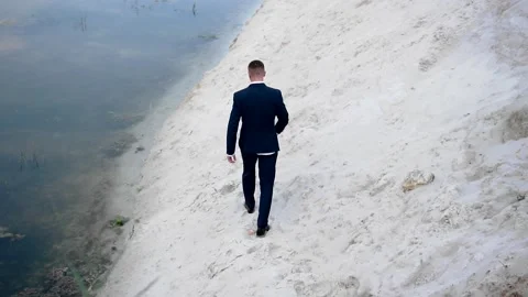 A tired guy in a jacket walks along the sandy shore of the pond Stock Footage 139229866