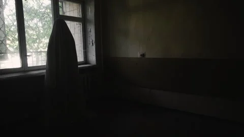 Tired halloween ghost in white sheet standing near window in dark room. Happy Stock Footage 314671082