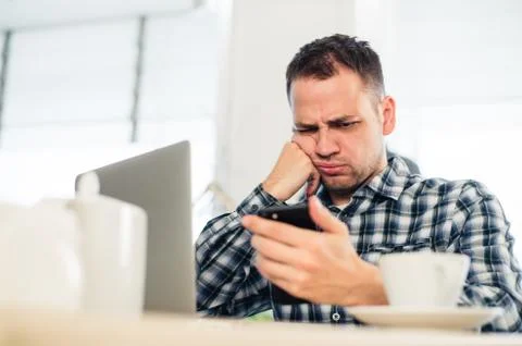 Tired handsome young freelancer is using a mobile phone while working at the Stock Photos
