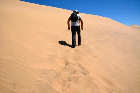 A tired man with a backpack on his back and wearing a cap climbs a desert sand Stock Photos