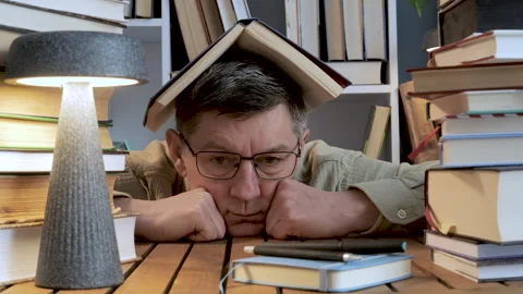 Tired man with book on head surrounded by stacks of books Stock Footage 336046214