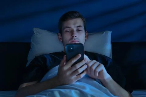 Tired Man Browsing Smartphone in Bed at Night 库存照片