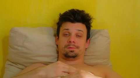 Tired man with hangover wakes up in bed looking at camera, portrait closeup. Stock Footage 128705331