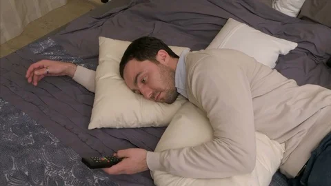 Tired man lays down on a bed to rest and uses a remote control to watch Vídeo Stock 74659559