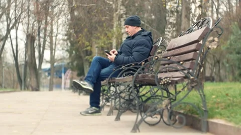 Tired man sits on a bench in a cold city alley and searches for information  Видео 317190988