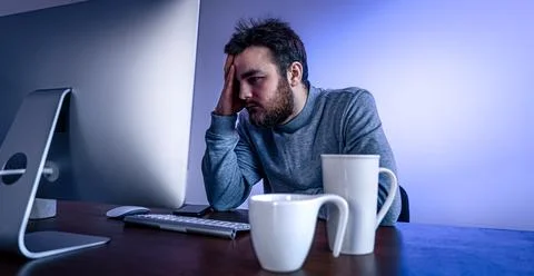 Tired man sits in front of a computer with a cup of coffee, colored lighting. Stock Photos
