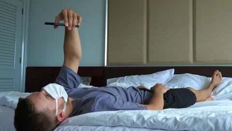 Tired man in the white face mask lying on the bed and checking in quarantine app Stock Footage 166304119