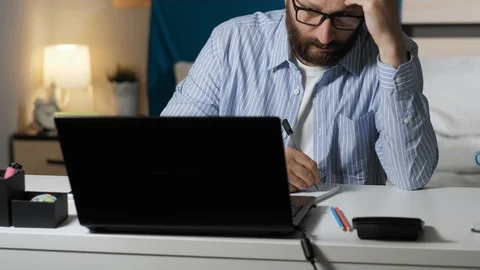 Tired man works at computer and writes notes in notebook. Bearded guy sits at Stock Footage 129379791