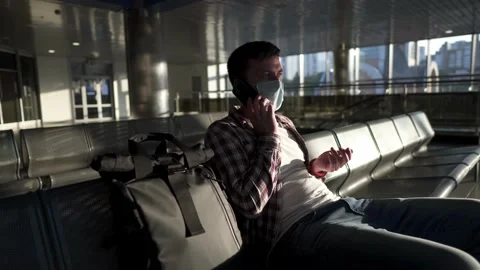 Tired, masked passenger sits in empty airport terminal, talking on phone with Vidéo 152521598