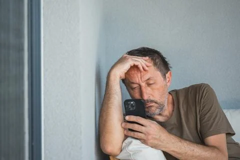 Tired middle-aged man leaning against wall using smartphone looking stresse.. Stock Photos