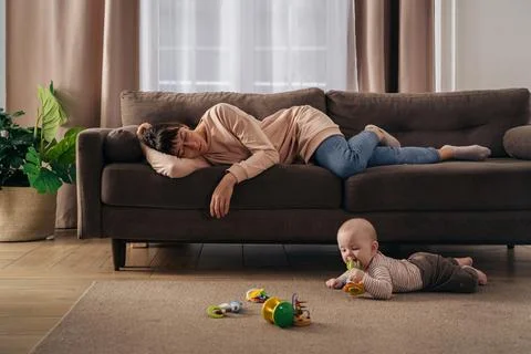 Tired mother sleeping on sofa while her toddler baby playing on floor Stock Photos