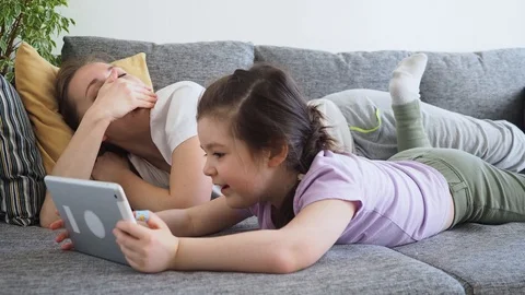 Tired mother sleeps while her small daughter using digital tablet on home's sofa Stock Footage 129478680