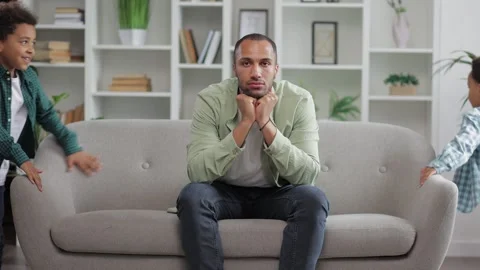 Tired parent father sitting on sofa while boys hyperactive kids are running a Stock Footage 253587630