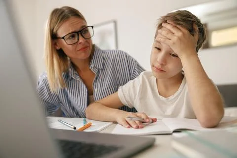 Tired sad kid sits at table with laptop and textbooks while mom help him with 스톡 사진