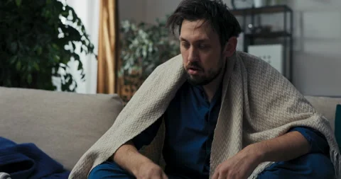 Tired sick bearded middle-aged man sitting on sofa in blue pajamas. Man sprays Видео 218331640