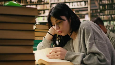 Tired student reading at busy library table with stacks of books and green lamps Stock Footage 336359211