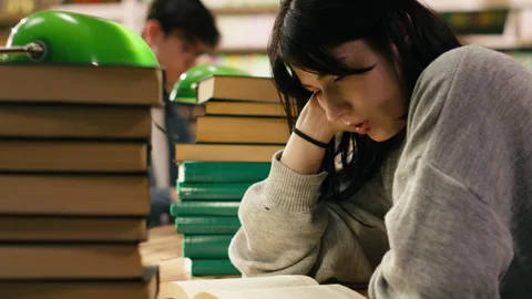 Tired student reading at busy library table with stacks of books and green lamps Stock Footage 336361093